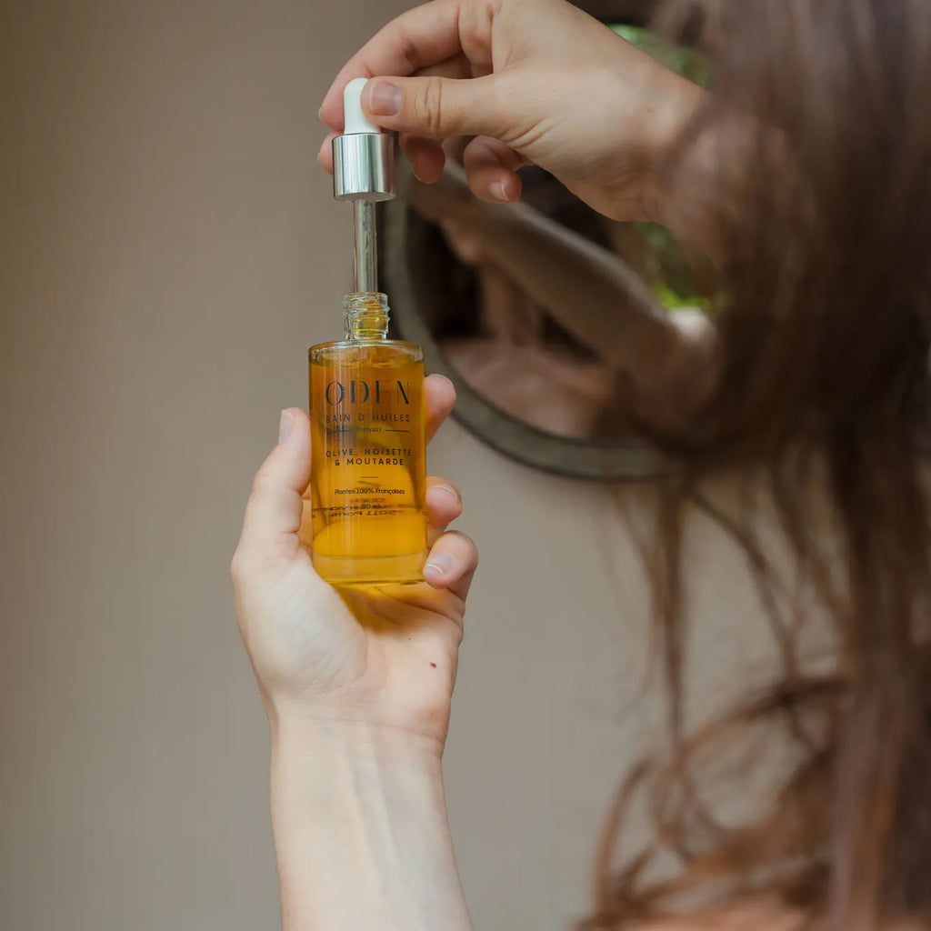 Person holding a bottle of Oden hair oil with a dropper, blurred background